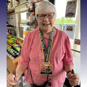 Edna Siniff stands smiling at the camera inside the A-1 Archery store in her hometown. A gold medal from the 2023 National Senior Games hands around her neck.