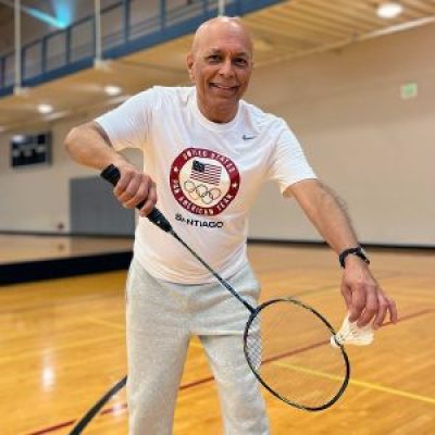 Dr. Rajeev Trehan stands in an indoor gym space. He is holding a badminton racket and shuttlecock while smiling at the camera.